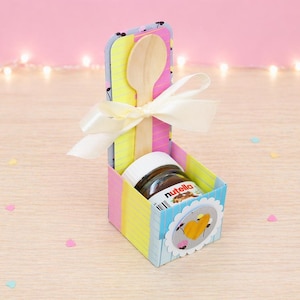 Op de afbeelding: Een kleurrijke geschenkdoos met een pot Nutella, een houten lepel en een wit lint. De doos is versierd met pastelkleuren en een decoratieve sticker. De achtergrond is lichtroze met feeërieke lichtjes.