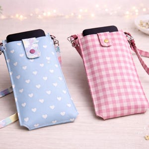 Puede incluir: Dos fundas verticales para teléfono con correas para el hombro. Una es azul claro con corazones blancos, la otra es rosa a cuadros. Ambas tienen un cierre de solapa con un botón. Las correas son de diferentes colores.