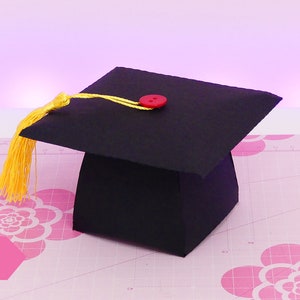 Può includere: Un cappello di laurea nero di carta con un fiocco giallo e un bottone rosso in cima. Il cappello è su un tappetino da taglio rosa e bianco con un motivo floreale.
