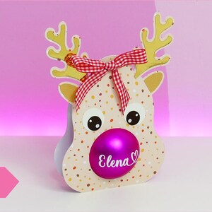 Könnte beinhalten: Ein dekorativer Rentier-Schmuck mit einer rosa Weihnachtskugel als Nase. Das Rentier hat große Augen, gelbe Geweihe und eine rot-weiß karierte Schleife. Der Körper des Rentiers ist beige mit bunten Punkten. Der Text "Elena" befindet sich auf der Kugel.