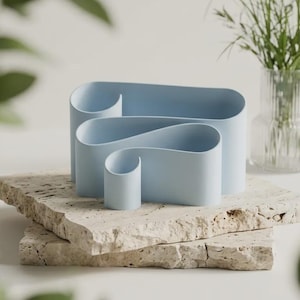 May include: A light blue desk organizer featuring a continuous, ribbon-like structure that creates three distinct compartments of varying heights. The object has a smooth, matte finish and a curved, fluid design that loops to form open storage spaces.
