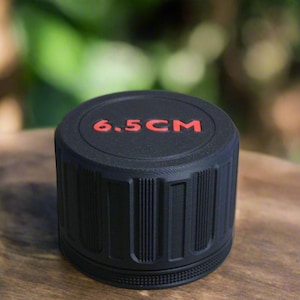 May include: A black cylindrical object with the text "6.5CM" in red on the top. The object has vertical grooves around the sides and sits on a wooden surface. The background is blurred green.