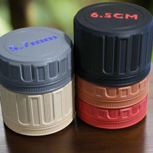 May include: Two cylindrical containers with textured sides and screw-on lids. One is gray and beige, the other is black, orange, and red. The black lid reads "6.5 CM" in red. The gray container has blue text on the lid.