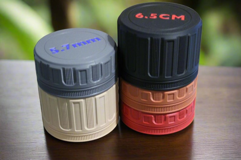 May include: Two cylindrical containers with textured sides and screw-on lids. One is gray and beige, the other is black, orange, and red. The black lid reads "6.5 CM" in red. The gray container has blue text on the lid.