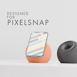 May include: A smartphone rests on an orange, spherical stand next to a gray, spherical speaker. The phone's screen displays a colorful pattern. Text on the image reads "DESIGNED FOR PIXELSNAP."