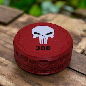 May include: A circular red grinder featuring a white skull graphic on the top lid and the number 380 printed in black below it. The exterior of the device has a textured, ridged grip pattern around the side, with a matte finish on the surface.