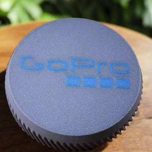 Pu&ograve; includere: Un tappo per obiettivo per fotocamera GoPro rotondo grigio con il logo GoPro in blu.