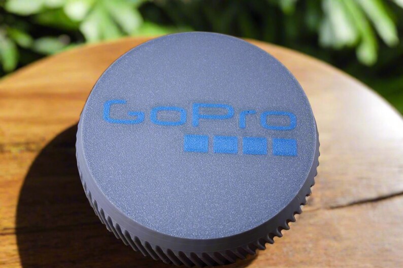 Pu&ograve; includere: Un tappo per obiettivo per fotocamera GoPro rotondo grigio con il logo GoPro in blu.