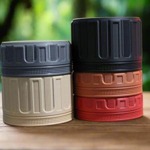 May include: A collection of cylindrical containers in various colors, including tan, black, red, and brown. Each container has a ribbed design on the lid and body. The containers are stacked and arranged on a wooden surface.