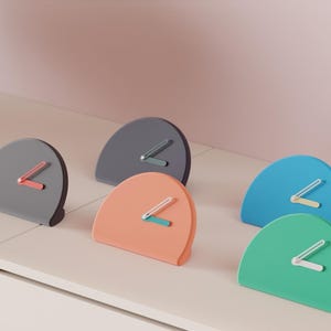 May include: A collection of modern, semi-circular clocks in various colors, including gray, coral, blue, and green. Each clock features a contrasting color for the hands, adding a pop of color to the minimalist design. The clocks are displayed on a white surface.
