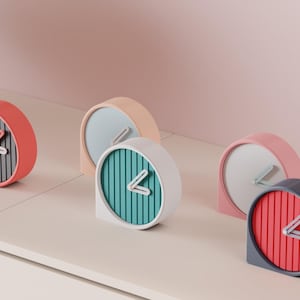 May include: A collection of modern, minimalist clocks in various pastel colors. Each clock has a rounded shape with a striped face and a single hand. The clocks are arranged on a light-colored surface against a pink background.