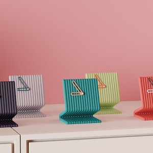 May include: A collection of modern desk clocks in various colors, including purple, lavender, teal, green, and red. Each clock has a unique, ribbed design and a minimalist clock face with a single hand. The clocks are displayed on a white surface against a pink background.