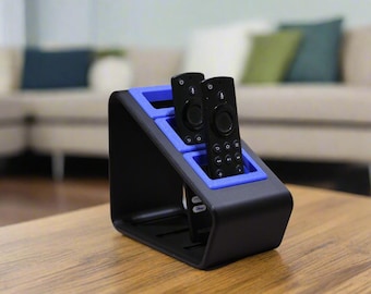 May include: A black and blue tiered remote control holder with a slanted design. The stand features three distinct slots, with two black remote controls inserted into the top and middle sections, and a third remote partially visible in the bottom slot.