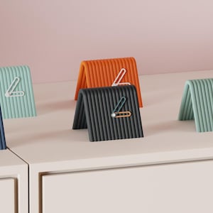May include: A collection of colorful, folded desk organizers in various shades, including teal, navy, orange, and black. Each organizer features a unique paperclip-style design in contrasting colors. The organizers are arranged on a white surface against a pink backdrop.