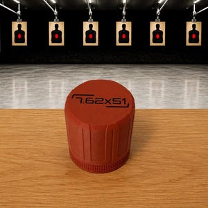 May include: A cylindrical red cap with vertical ridges and a textured base, featuring the black text 7.62x51 printed on the top surface. The object is positioned in the foreground against a blurred background of an indoor shooting range.