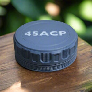 May include: A gray, cylindrical object with the text "45ACP" on top. The object has a textured, ridged design around the sides. It sits on a wooden surface, with a blurred green background.