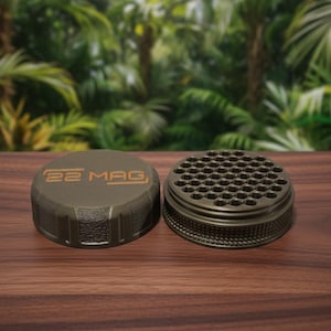 May include: Two olive-green metal containers sit on a wooden surface. One is a lid with the text "22 MAG" in orange. The other is a cylindrical container with numerous small holes. The background is a blurred view of green foliage.
