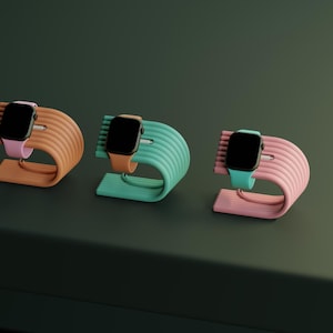 May include: Three colorful watch stands in peach, teal, and pink, each holding a black smartwatch with a matching band. The stands have a curved, ribbed design and are displayed on a dark green surface, showcasing a modern aesthetic.
