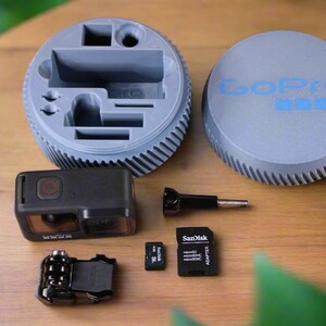 Pu&ograve; includere: Una custodia grigia per fotocamera GoPro con un vano per gli accessori. La custodia ha il logo GoPro sul coperchio. L'immagine mostra anche una fotocamera GoPro nera, un adattatore per schede di memoria nero, due schede di memoria e un accessorio nero.