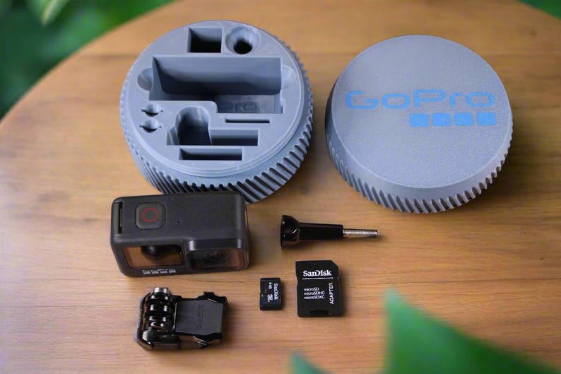 Pu&ograve; includere: Una custodia grigia per fotocamera GoPro con un vano per gli accessori. La custodia ha il logo GoPro sul coperchio. L'immagine mostra anche una fotocamera GoPro nera, un adattatore per schede di memoria nero, due schede di memoria e un accessorio nero.
