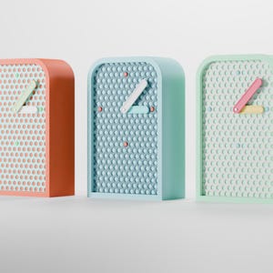 May include: Three colorful clocks in a row. The clocks have a rectangular shape with rounded corners. The clock faces are covered in small, circular indentations. The clock bodies are in shades of orange, blue, and green. The clock hands are white, pink, and yellow.