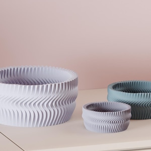 May include: Three decorative, round bowls in varying sizes and colors. The largest bowl is white, the medium bowl is a light purple, and the smallest bowl is a teal color. Each bowl has a wavy, textured design.
