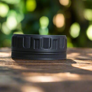 May include: A black, cylindrical object with a textured surface. The object has a series of vertical rectangular indentations around the top and a dotted pattern around the middle. It sits on a wooden surface with a blurred green background.