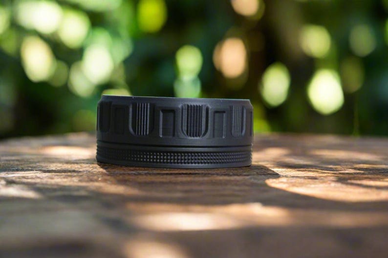 May include: A black, cylindrical object with a textured surface. The object has a series of vertical rectangular indentations around the top and a dotted pattern around the middle. It sits on a wooden surface with a blurred green background.