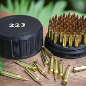 May include: A collection of .223 caliber bullets, with a black container labeled "223". The bullets are gold-colored with pointed tips, arranged in a black holder and scattered on a wooden surface.