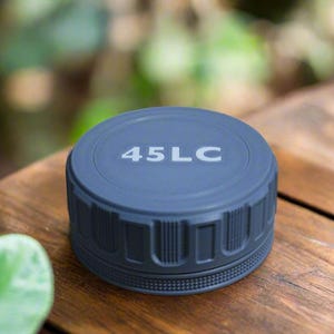 May include: A dark gray, cylindrical object with the text "45LC" in white. The object has a ridged, textured design around its sides and sits on a wooden surface. The background is blurred, with green foliage visible.