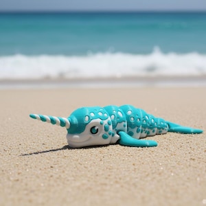 May include: A turquoise and white toy narwhal with a spiraled horn rests on a sandy beach. The toy has a segmented body with white spots and small fins. The background shows the ocean and sky.