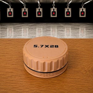 May include: A tan, circular, flat-topped container lid with vertical ridges along the side and a textured grip band at the base. The top surface features the black printed text 5.7X28, indicating the caliber for which this item is designed.