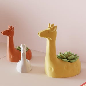 May include: Three ceramic animal planters: a giraffe in yellow, a giraffe in orange, and a white llama. The giraffe planters hold green succulents. The planters are on a white surface against a pink background.