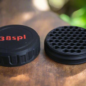 May include: Two black, circular objects sit on a wooden surface. One has the red text "38spl" on top. The other has a honeycomb pattern on top. Both have a ridged edge.