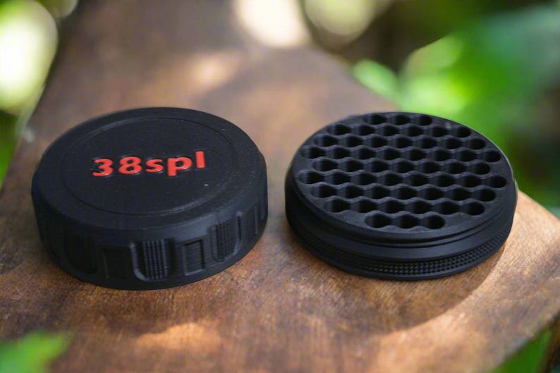 May include: Two black, circular objects sit on a wooden surface. One has the red text "38spl" on top. The other has a honeycomb pattern on top. Both have a ridged edge.