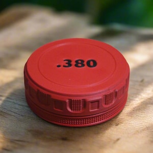 May include: A red, circular object with the text ".380" in black. The object has a textured edge and sits on a wooden surface. The background is blurred, suggesting an outdoor setting.