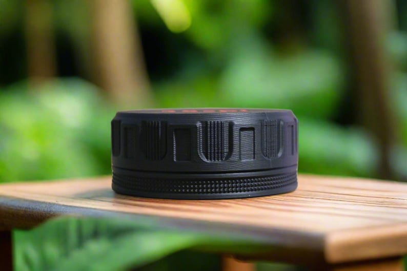 May include: A black, cylindrical object with a textured surface and a ridged design. The object is sitting on a wooden surface, with a blurred green background. The top of the object has a red detail.