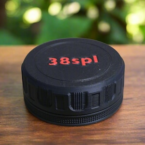 May include: A black, cylindrical object with the text "38spl" in red lettering on the top. The object has a textured surface and is sitting on a wooden surface. The background is blurred.