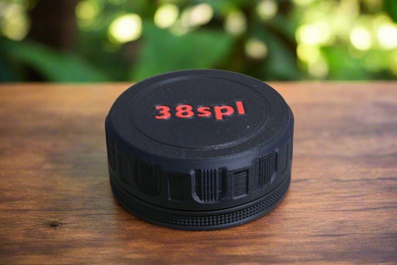 May include: A black, cylindrical object with the text "38spl" in red lettering on the top. The object has a textured surface and is sitting on a wooden surface. The background is blurred.