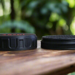 May include: Two black jar lids are displayed on a wooden surface. One lid has a textured, ridged design with red text, while the other has a circular, patterned surface. The lids are likely for food storage.