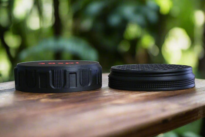 May include: Two black jar lids are displayed on a wooden surface. One lid has a textured, ridged design with red text, while the other has a circular, patterned surface. The lids are likely for food storage.