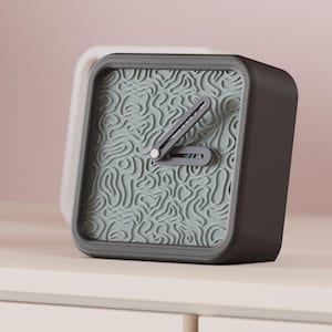 May include: A dark gray square alarm clock with a light gray face featuring a topographic pattern. The clock hands are gray. A white clock is partially visible behind the main clock. The clock is on a white surface.
