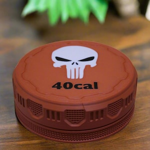 May include: A round, brown container with a white skull graphic and the text "40cal" in black. The container has a textured design around the sides and is sitting on a wooden surface. The product is likely a storage container.