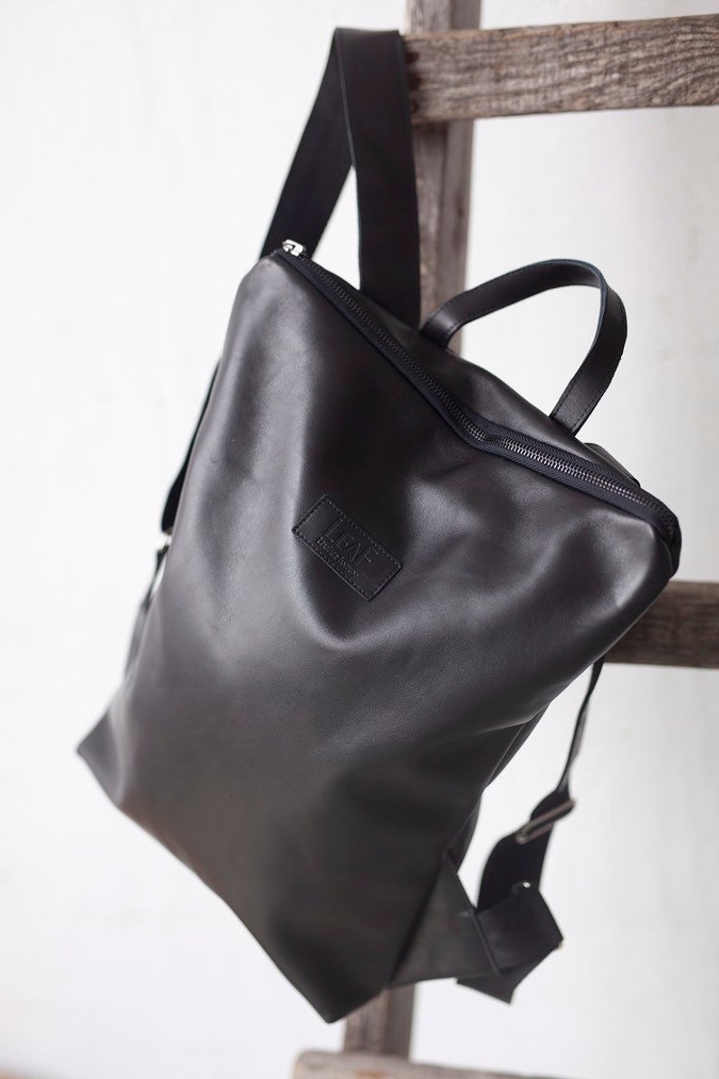 May include: A black leather backpack with a zipper closure and a small leather patch with the text "LEAF" on the front.