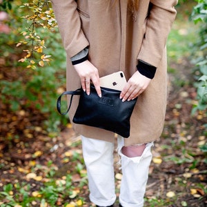 May include: A black leather clutch with a wrist strap. The clutch is closed and has a small rectangular label on the front.