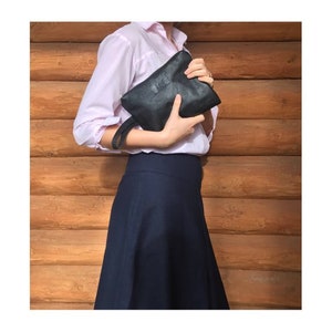 May include: A woman wearing a pink button-down shirt and a dark blue skirt is holding a black leather clutch. The clutch has a small silver logo on the front.
