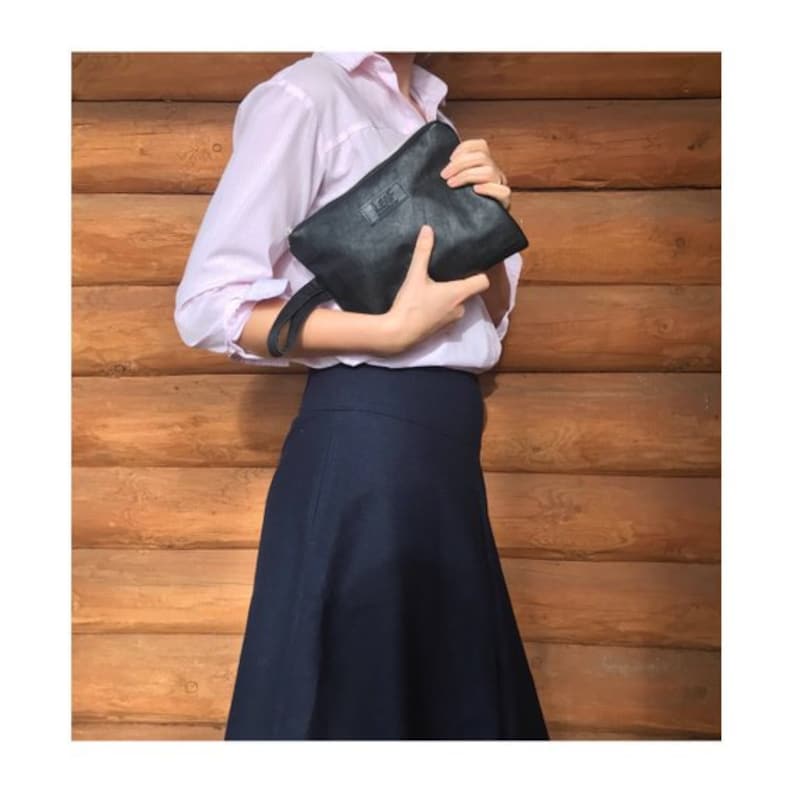 May include: A woman wearing a pink button-down shirt and a dark blue skirt is holding a black leather clutch. The clutch has a small silver logo on the front.