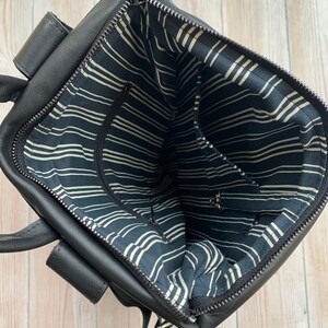 May include: Black leather backpack with a zippered top opening. The inside of the bag is lined with black and white striped fabric. The bag has two adjustable straps.