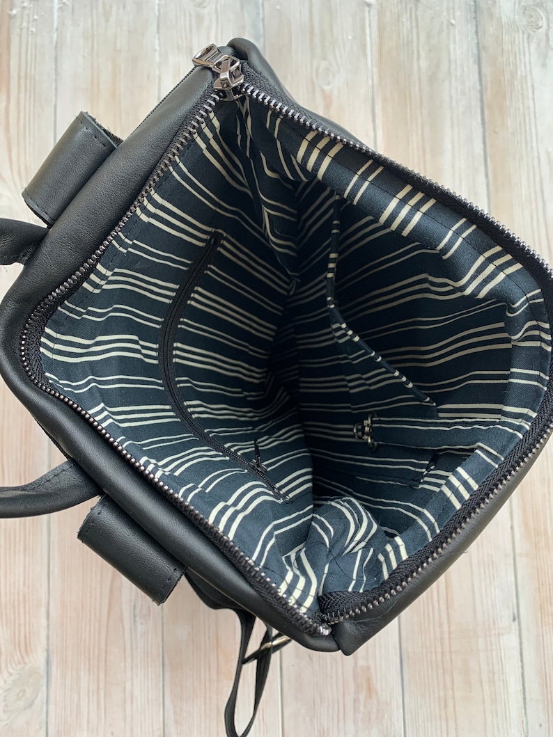 May include: Black leather backpack with a zippered top opening. The inside of the bag is lined with black and white striped fabric. The bag has two adjustable straps.