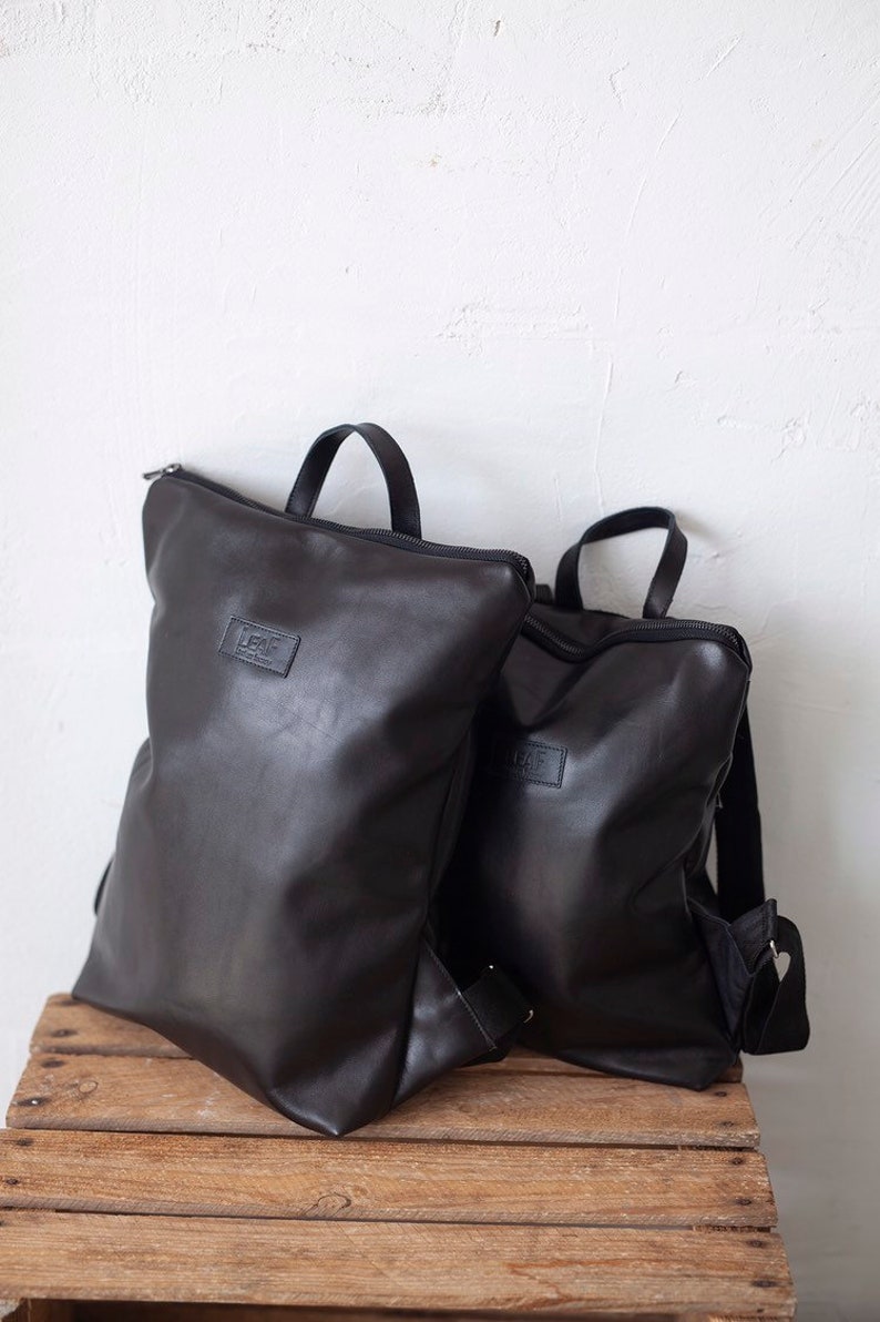 May include: Two black leather backpacks with zipper closures and adjustable straps. The larger backpack has the word "LEAF" printed on the front.